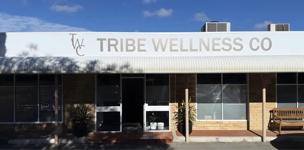 Exterior of Tribe Wellness Co building with signage, large windows, potted plants, and a wooden bench, under a bright blue sky