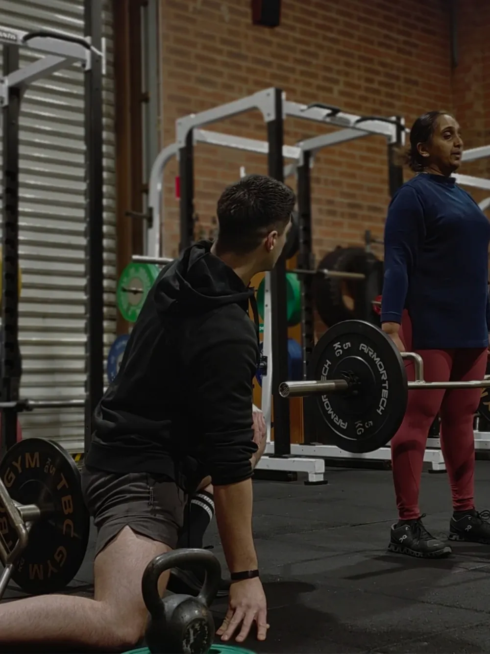Man kneeling on the gym floor with a kettlebell while a coach stands beside him